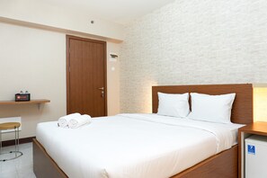 Quarto de hotel com cama de madeira, roupa de cama branca, uma pequena escrivaninha com cadeira, guarda-roupa de madeira e aquecedor fixado na parede.
