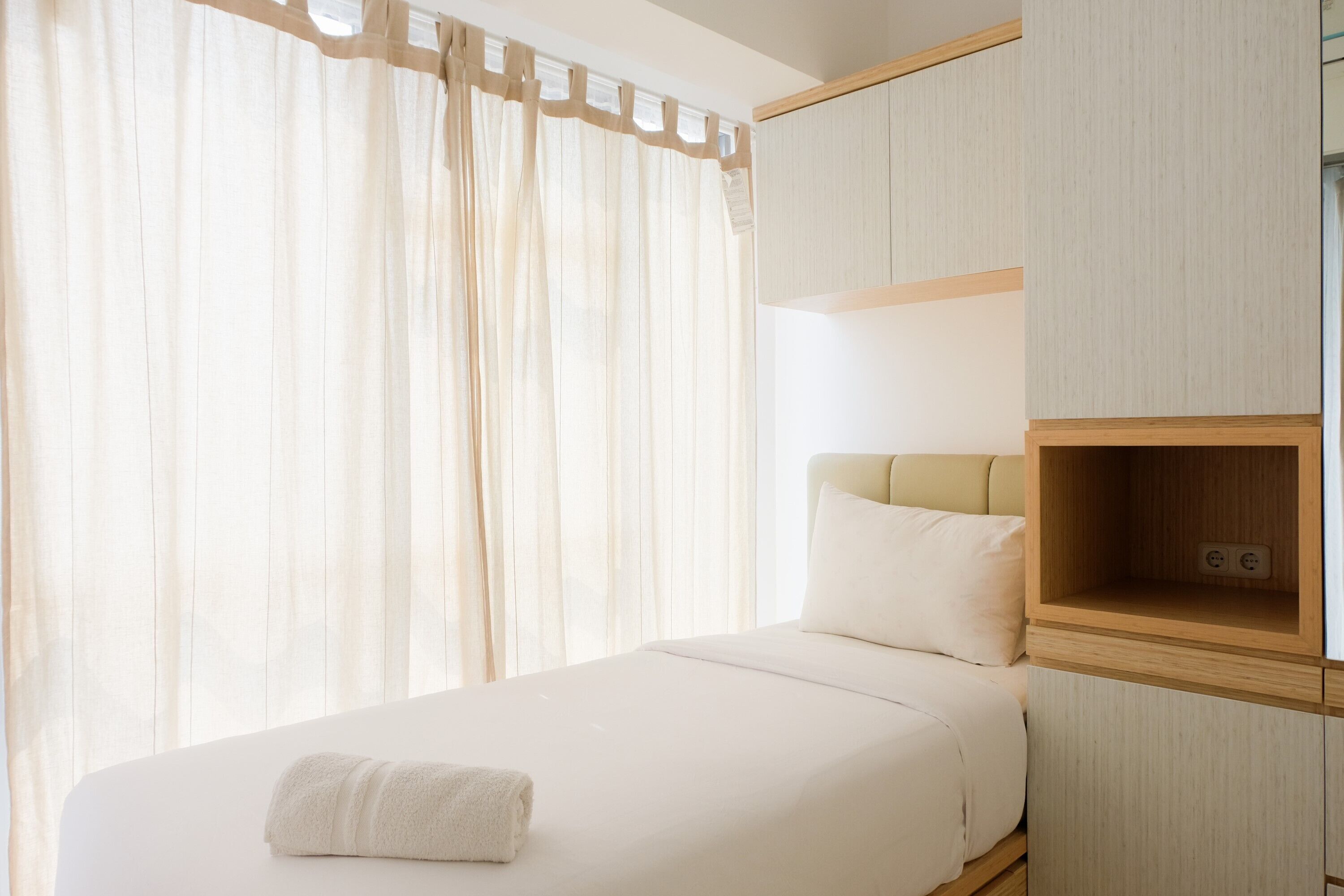 A neatly made bed with a white comforter and a folded white towel. There is a wooden headboard and a small wooden shelf with an electrical outlet.