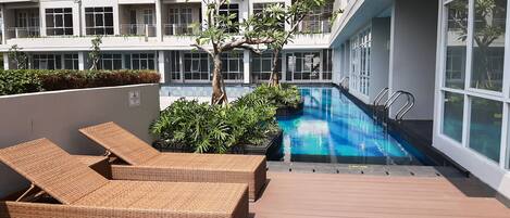 A rooftop pool area with two wicker lounge chairs, a small tree, and a modern building in the background.