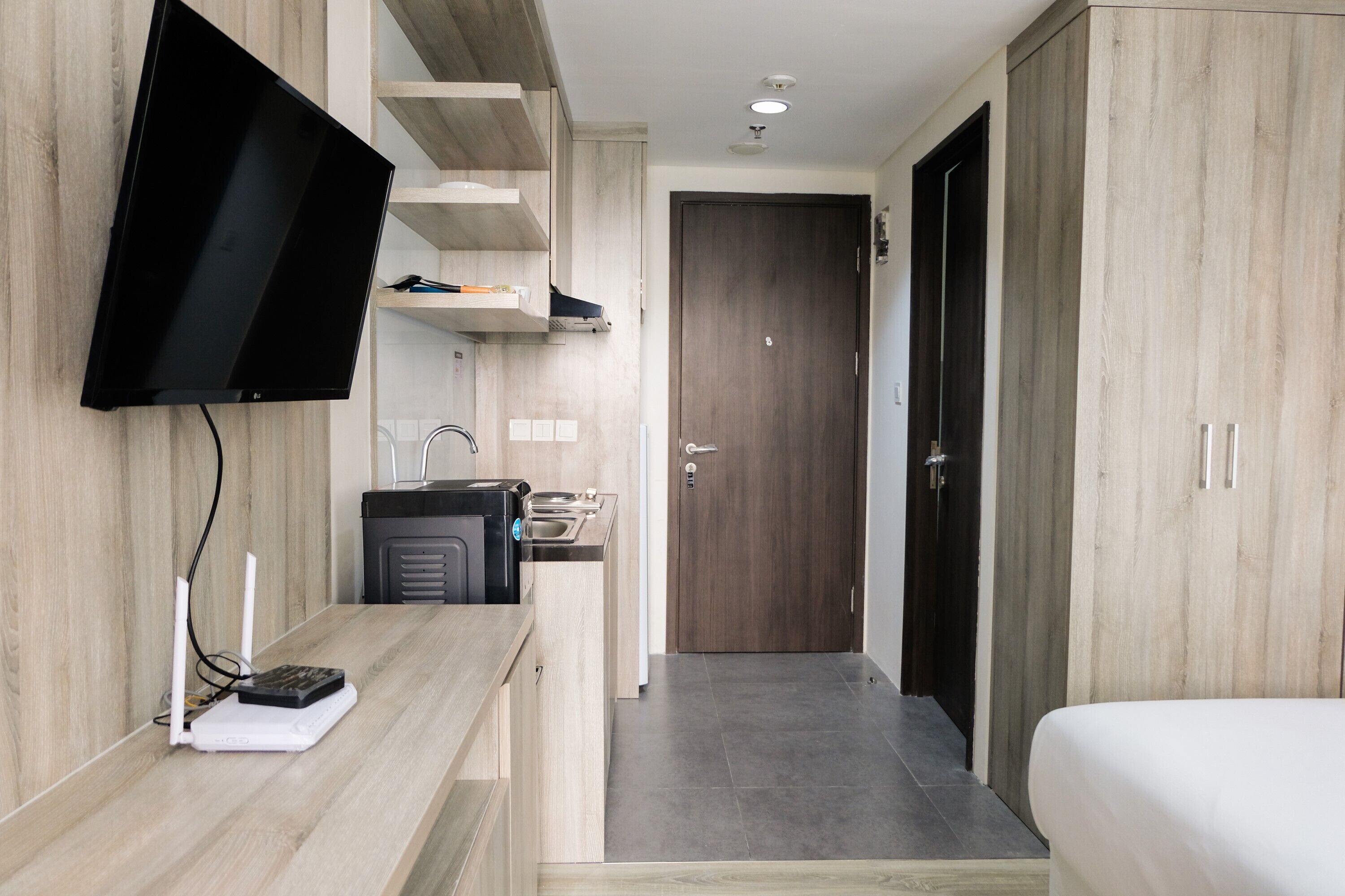 A modern hotel room with a flat-screen TV mounted on the wall, a kitchenette with a sink and a small refrigerator, and a wooden wardrobe with a door.