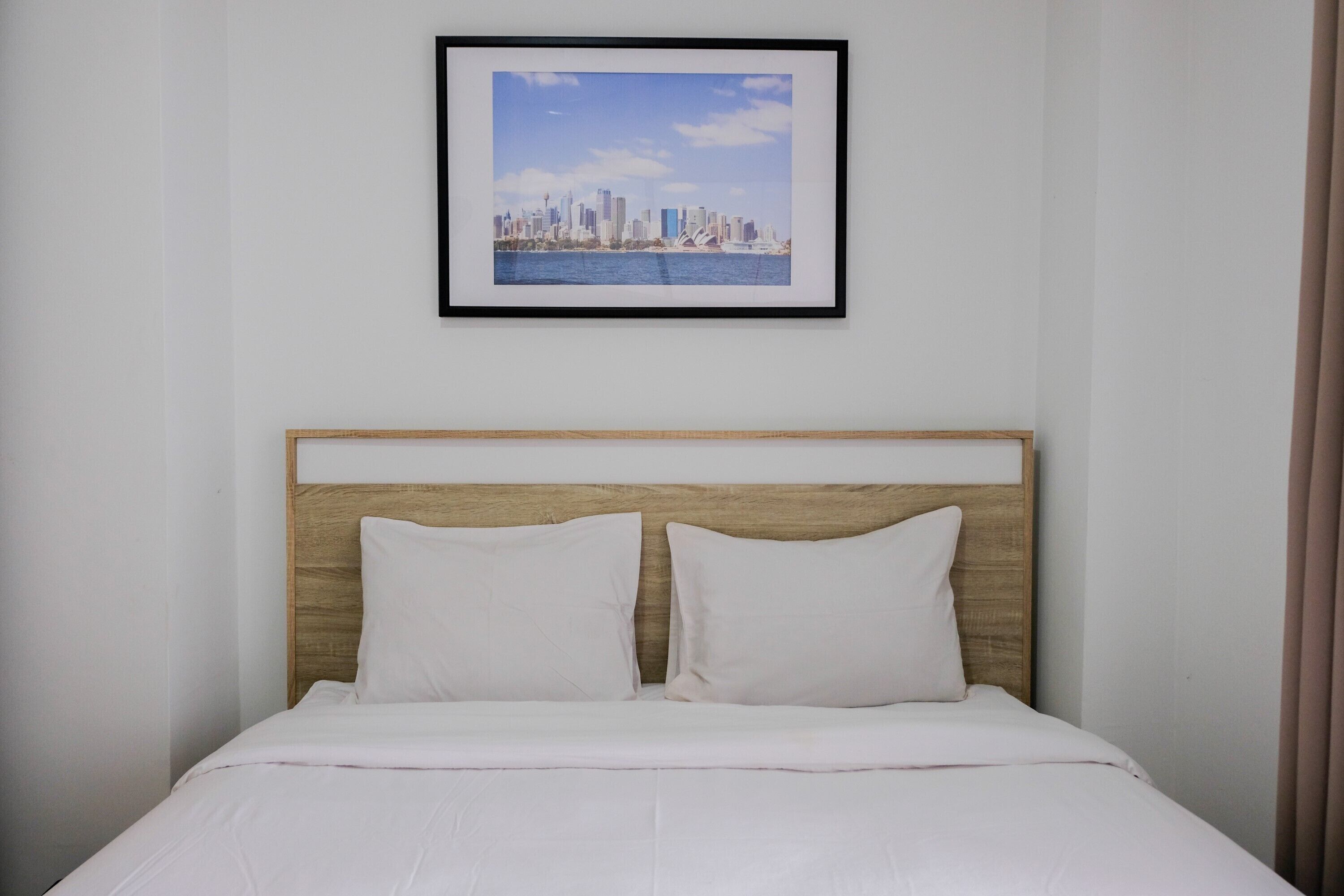 A neatly made bed with a wooden headboard, white bedding, and a framed cityscape picture above it.