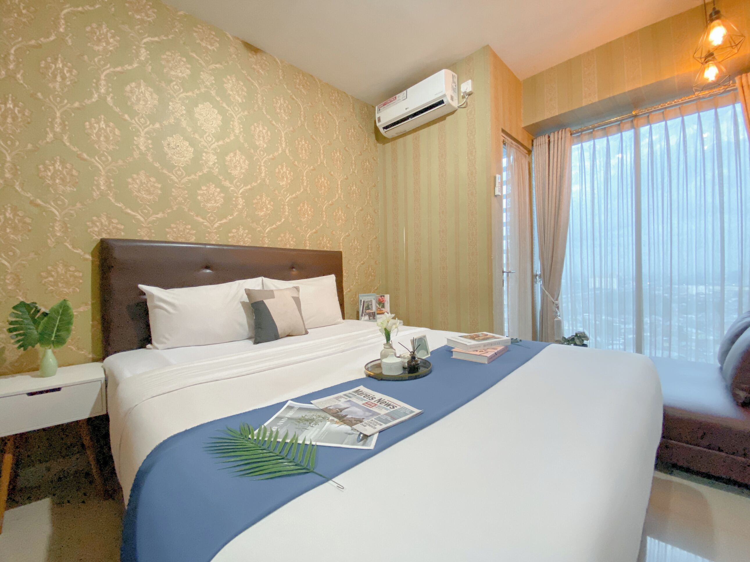 A hotel room with a bed, a nightstand, an air conditioning unit, a window with curtains, and a view of the city.