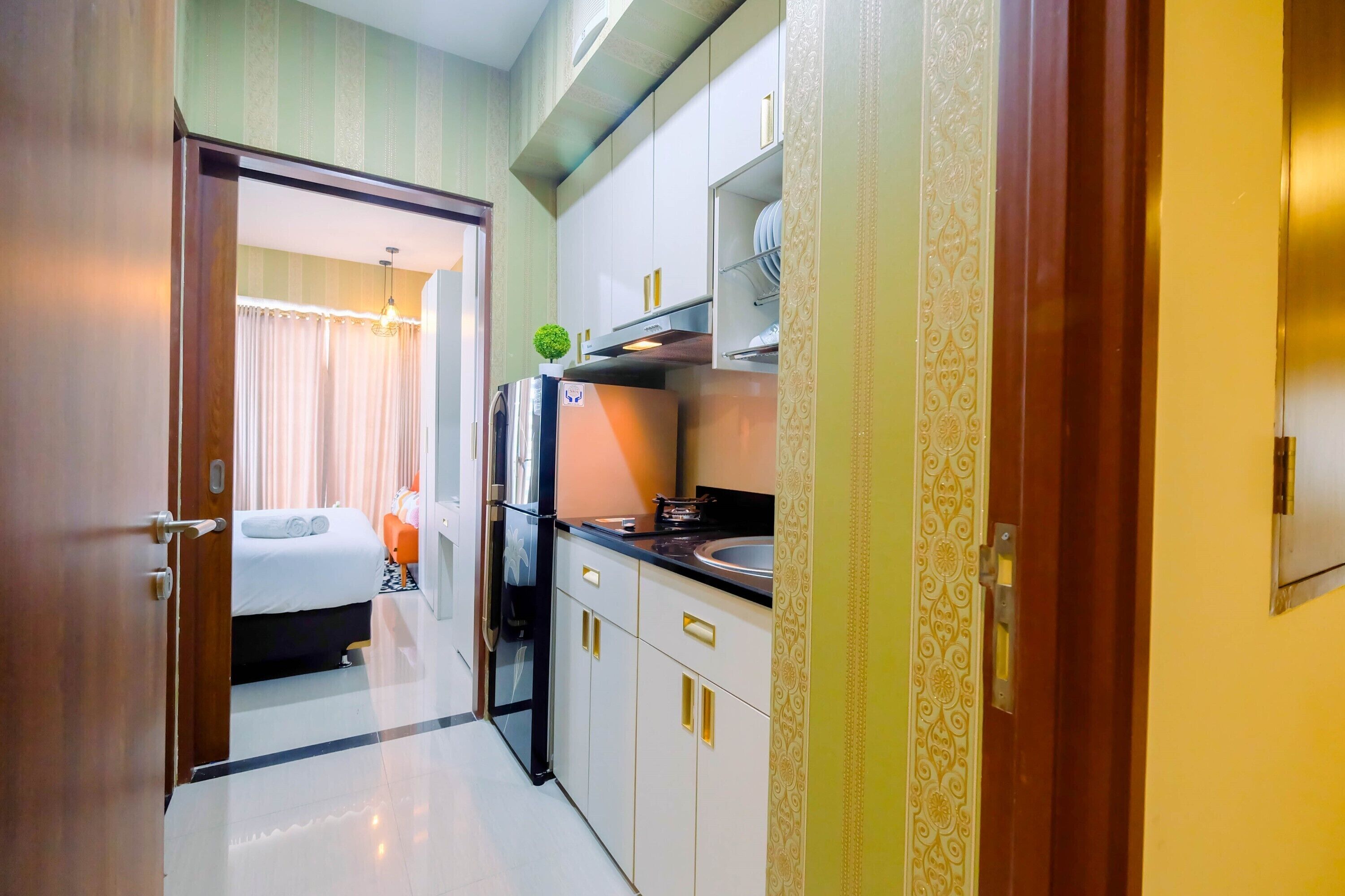 Un couloir étroit mène à une chambre à coucher équipée d’un lit et d’une table de chevet, à une cuisine avec des meubles blancs et un réfrigérateur, et à une porte ornée de motifs décoratifs.