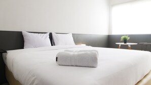 A neatly made bed with white linens and a folded towel, a bedside table with a lamp, and a small potted plant on a round table. - Comfy Studio Room @ Oasis Apartment (Cikarang)