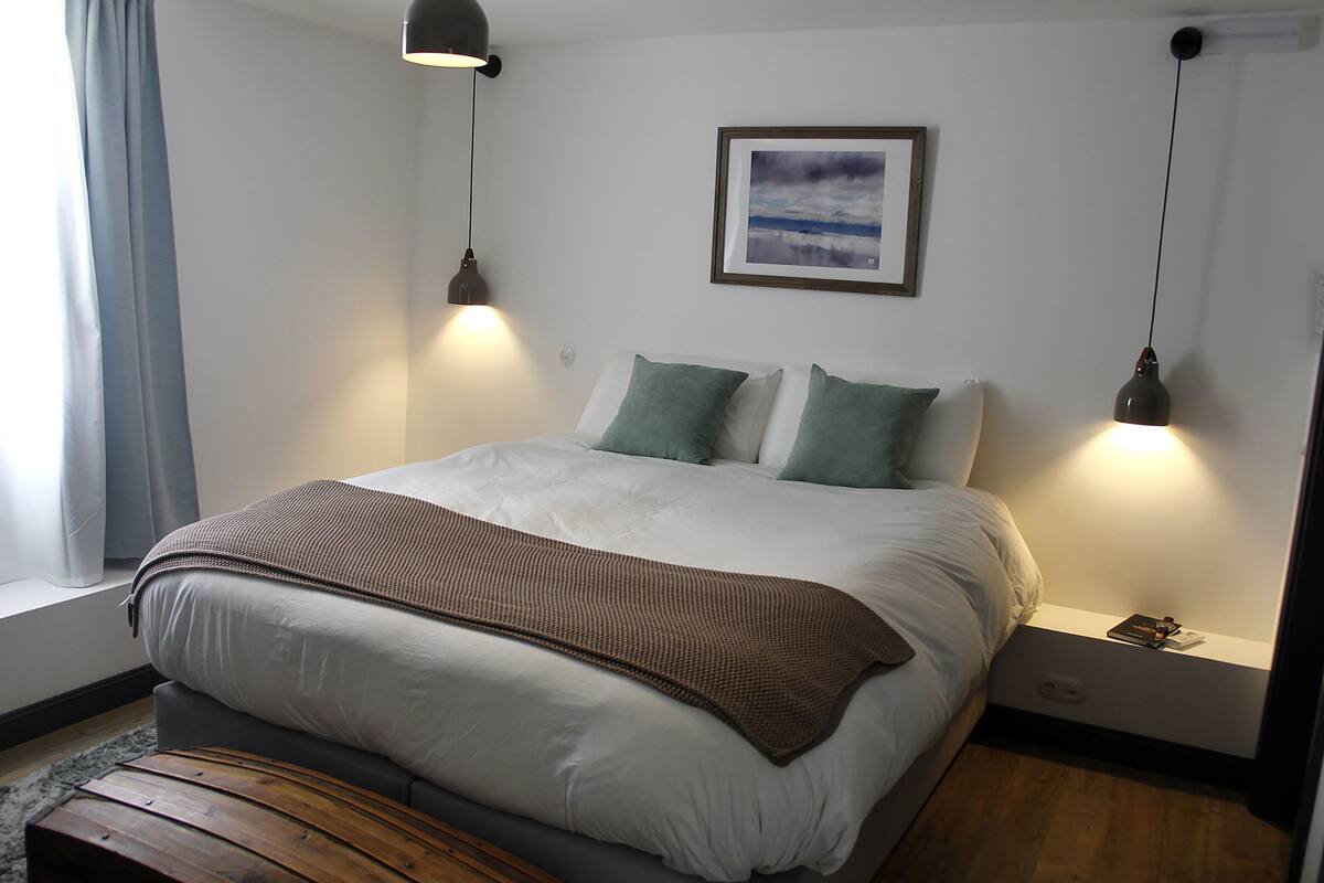 Un lit bien fait, avec des oreillers et une couverture, une table de chevet avec une lampe et un tableau encadré au mur.