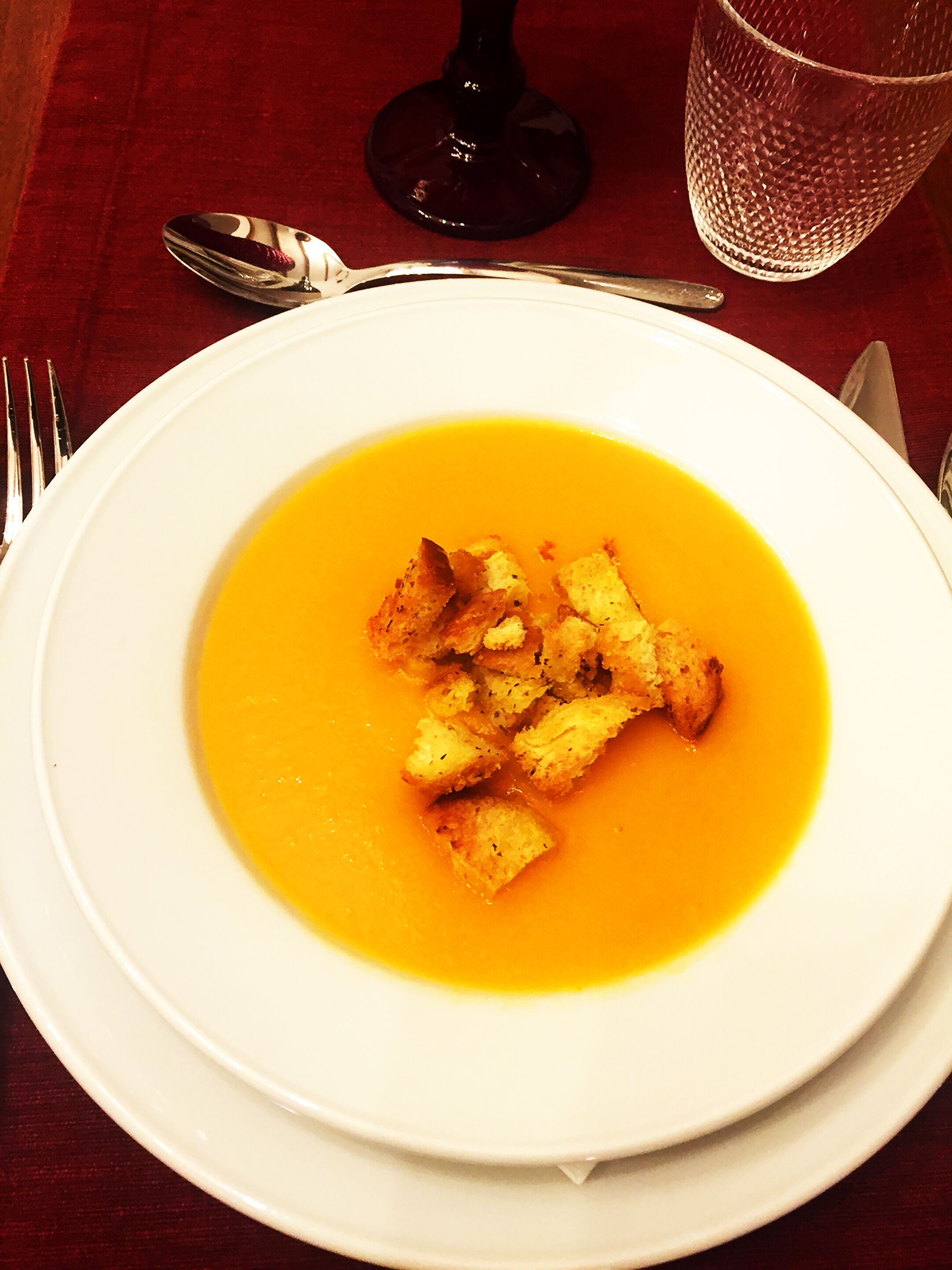 A white plate with a bowl of soup and croutons on a table.