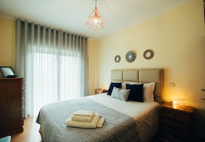 A neatly made bed with a headboard, a bedside table with a lamp, and a window with curtains.
