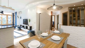 Een moderne eetruimte met een houten tafel voor vier personen, een televisie op de achtergrond en een stadszicht door grote ramen.