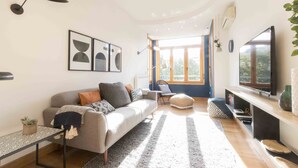 Een moderne woonkamer met een bank, een salontafel en een televisie. Er hangen ingelijste foto’s aan de muur en een raam zorgt voor natuurlijk licht.