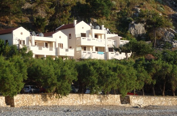 A multi-story building with balconies, surrounded by trees and rocky terrain. - Ravdoucha Beach Studios (Platanias)