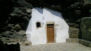 Un edificio blanco con puerta de madera y una ventana pequeña, rodeado de muros de piedra y un camino empedrado.