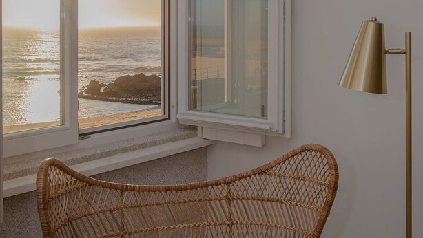 A room with a wicker chair near a window overlooking the ocean, a floor lamp, and a patterned throw on the chair. - Liiiving in Porto| Luxury Beachfront Apartment III (Porto)