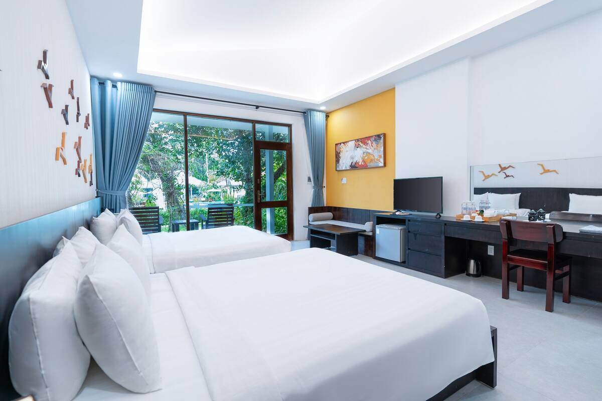 A hotel room with two beds, a desk, a chair, a TV, and a window with a view of greenery.