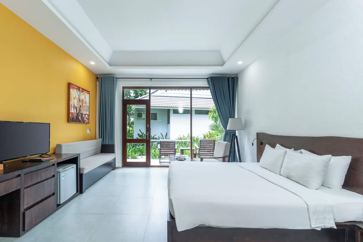 A hotel room with a bed, a TV, a desk, and a balcony with a view of greenery.