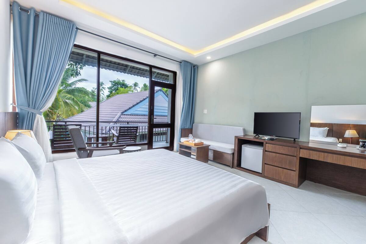 A hotel room with a large bed, a desk with a TV, a small kitchenette, and a balcony with a view of the outside.
