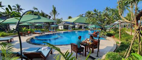A pool area with lounge chairs, umbrellas, and a dining table with people.