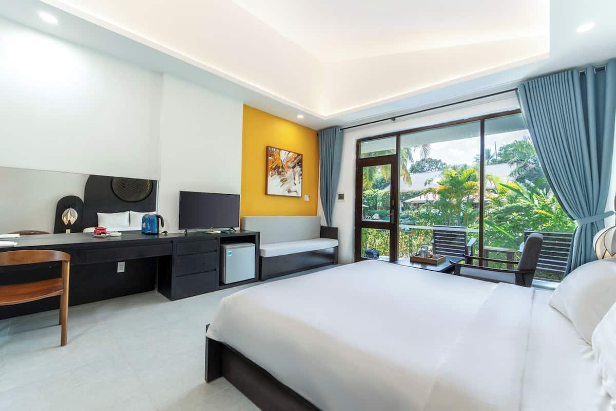 A hotel room with a large bed, a desk with a computer, a small fridge, and a view of greenery through the window.