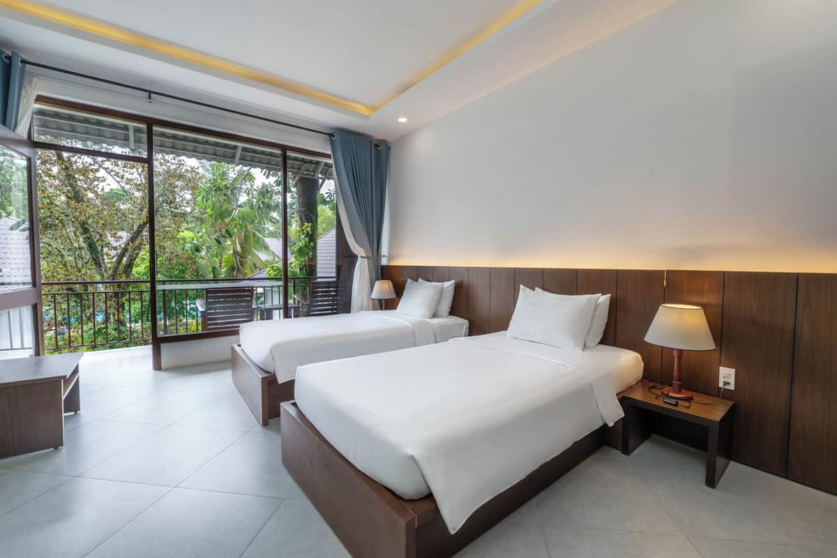 A hotel room with two beds, a balcony with a view of trees, and a desk with a lamp.