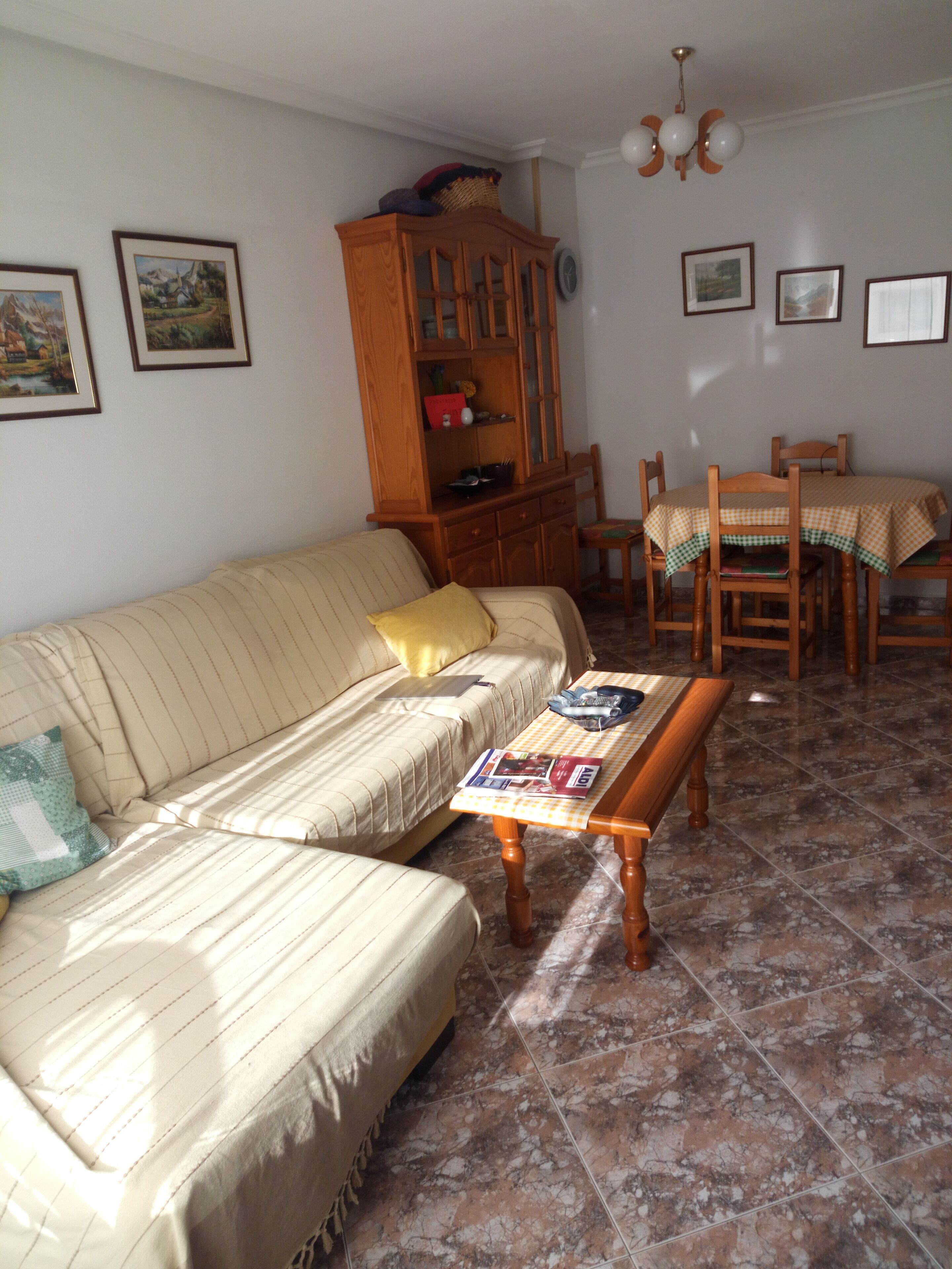 A living room with a sofa, a wooden cabinet, a dining table with chairs, and a tiled floor.