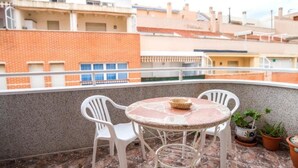 Un balcón con una mesa redonda y dos sillas, plantas en macetas y vistas de edificios al fondo.