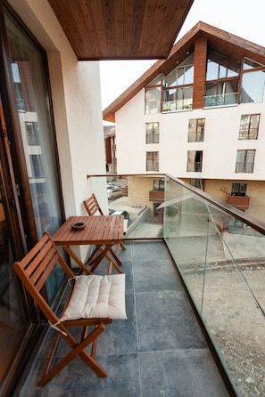 Ein Balkon mit Holztisch und -stühlen, einem Glasgeländer und Blick auf ein Gebäude mit zahlreichen Fenstern und Balkonen.
