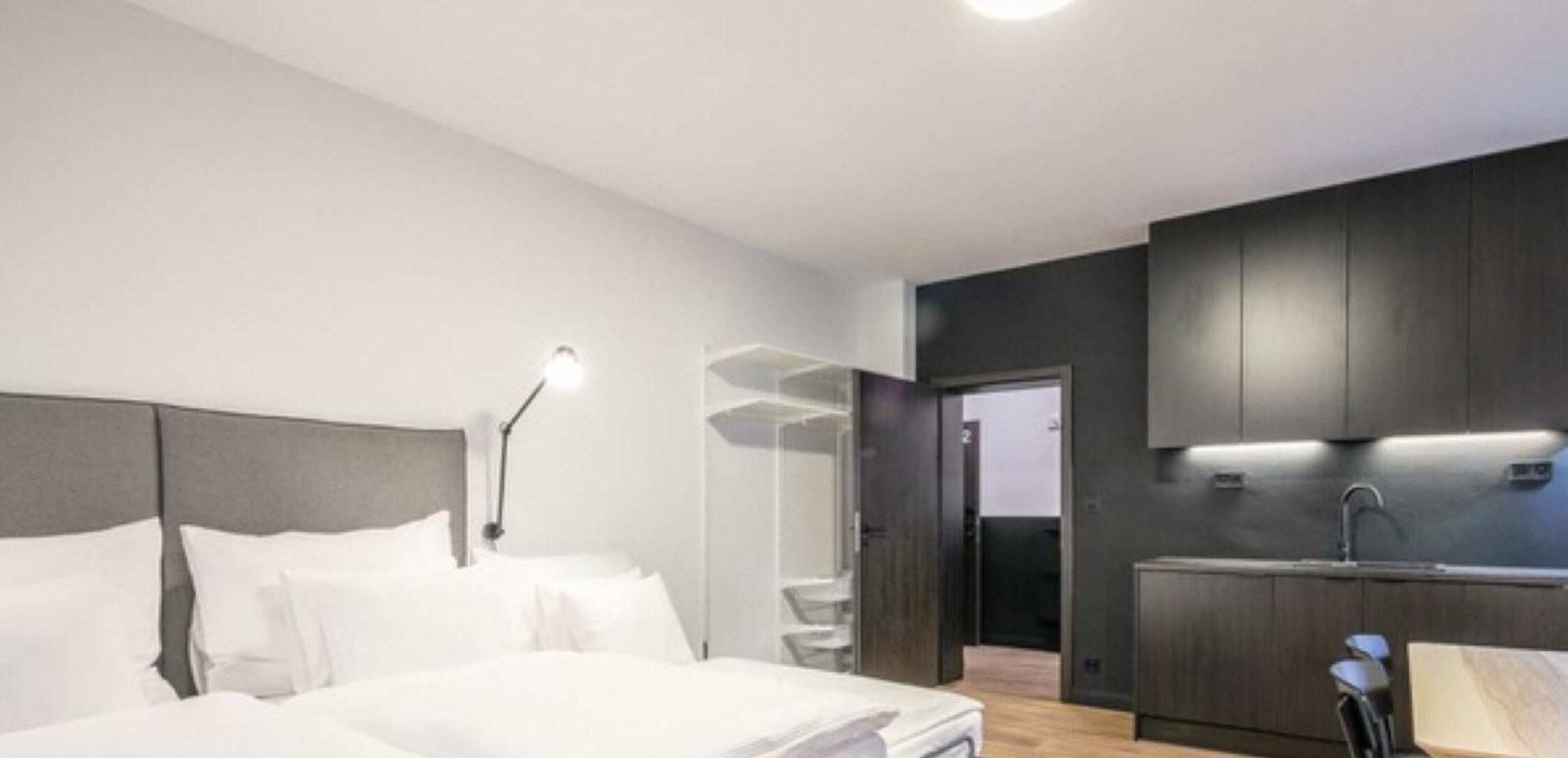 Una habitación de hotel moderna con una cama grande, mesita de noche y una zona de cocina integrada.