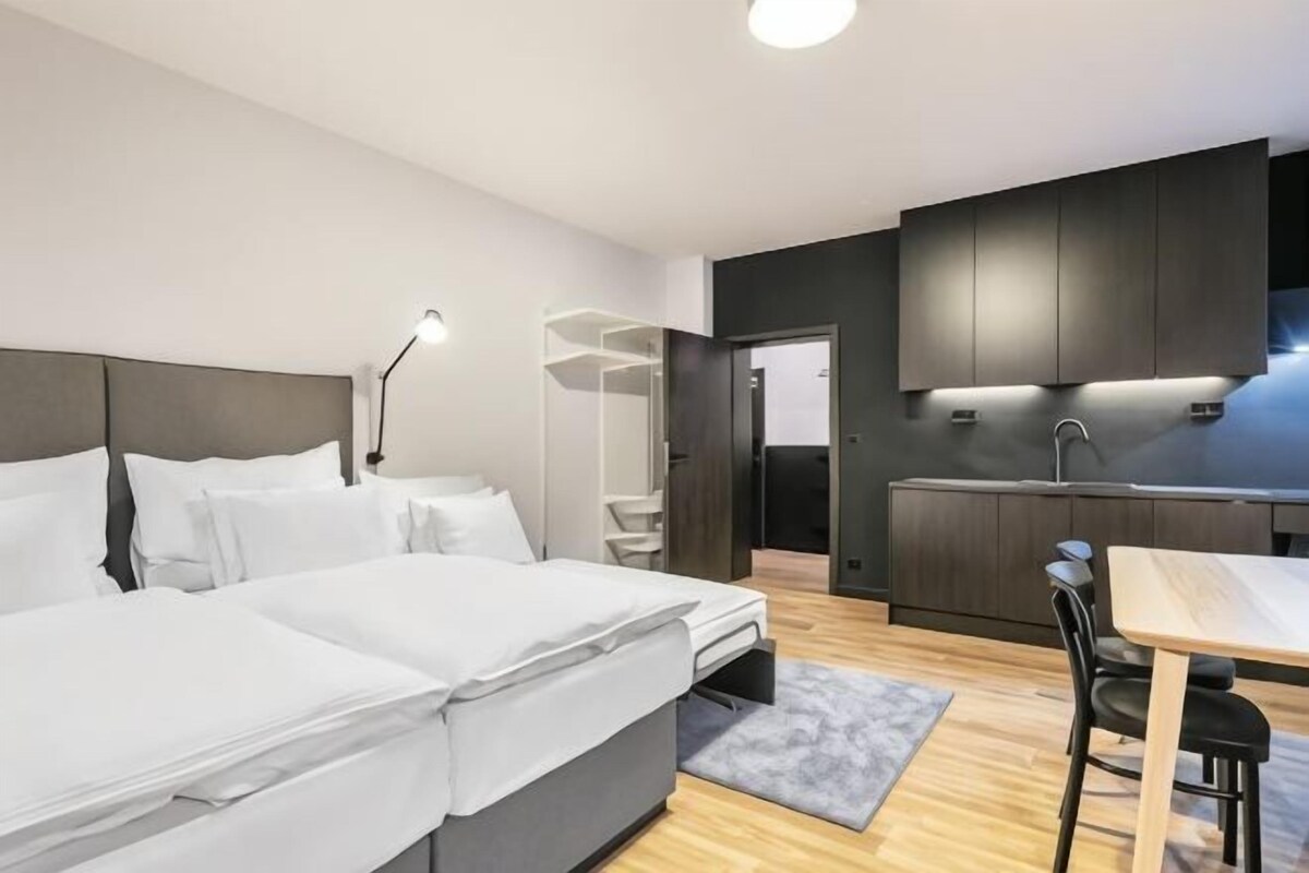 A modern hotel room with a bed, bedside table, desk, chair, and kitchenette.