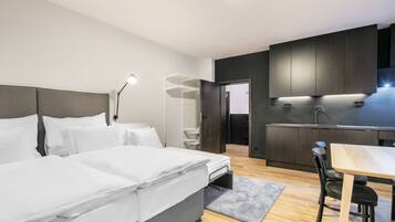 Une chambre d’hôtel moderne dotée d’un grand lit, d’une table de chevet et d’un bureau avec une chaise. Elle comprend également une kitchenette équipée d’un évier et de placards.