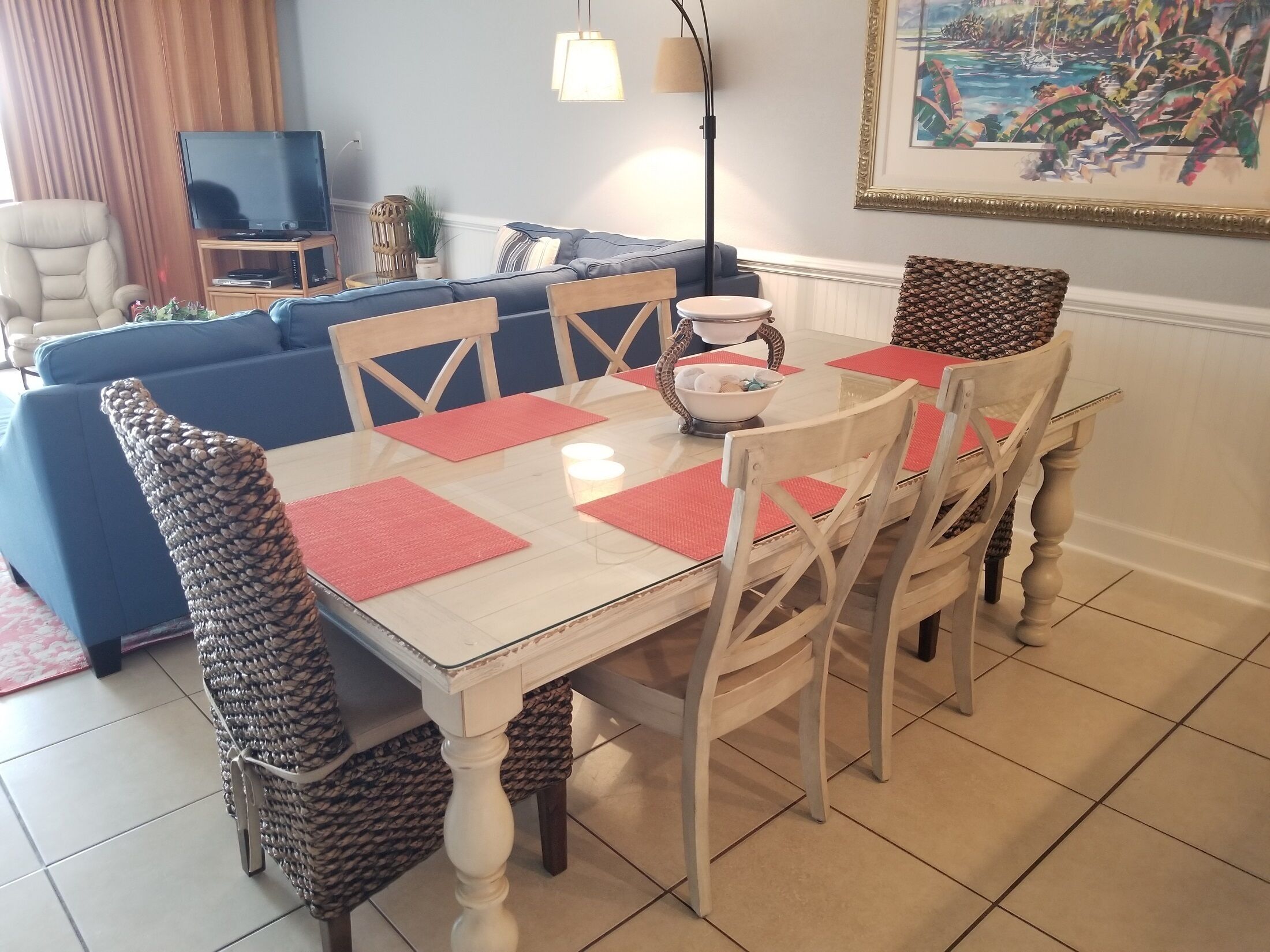 A dining area with a white table, red placemats, and wicker chairs. A blue sofa and a television are in the background.