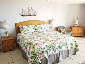 A bedroom with a large bed, a wicker headboard, a wall-mounted ship decoration, a small table, a chair, a dresser, and two bedside lamps. - Hibiscus B203 (St. Augustine)
