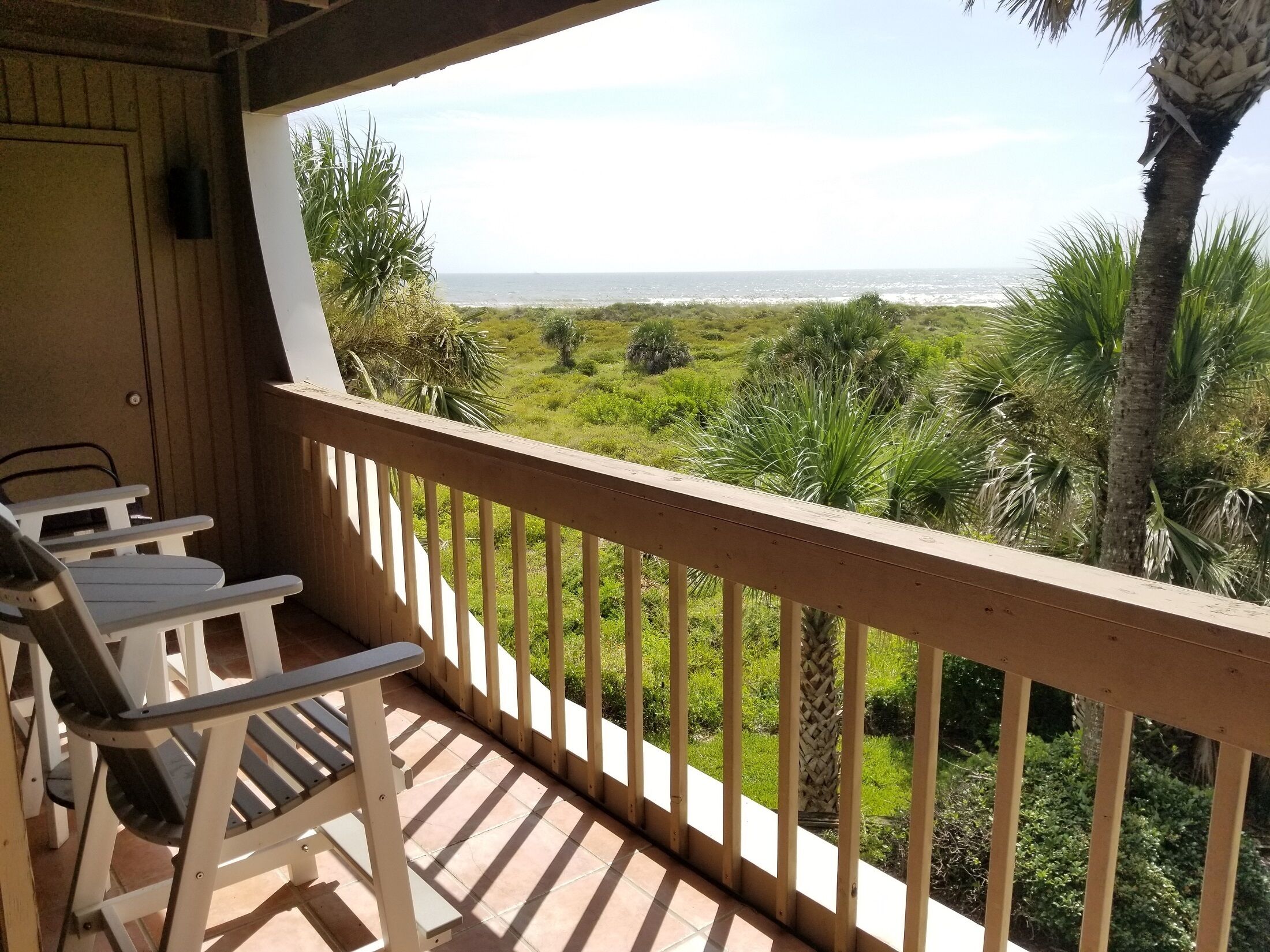 A wooden deck with a railing, white chairs, and a view of the ocean and greenery.