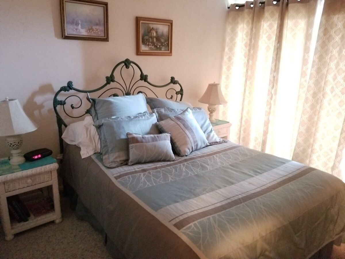Een net opgemaakt bed met een beddengoed met patroon en meerdere kussens, een nachtkastje met een lamp, en twee ingelijste foto’s aan de muur.