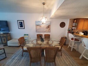 Un comedor con una mesa de vidrio rodeada de sillas de mimbre, un cuadro en la pared y una zona de cocina con armarios de madera y un reloj.