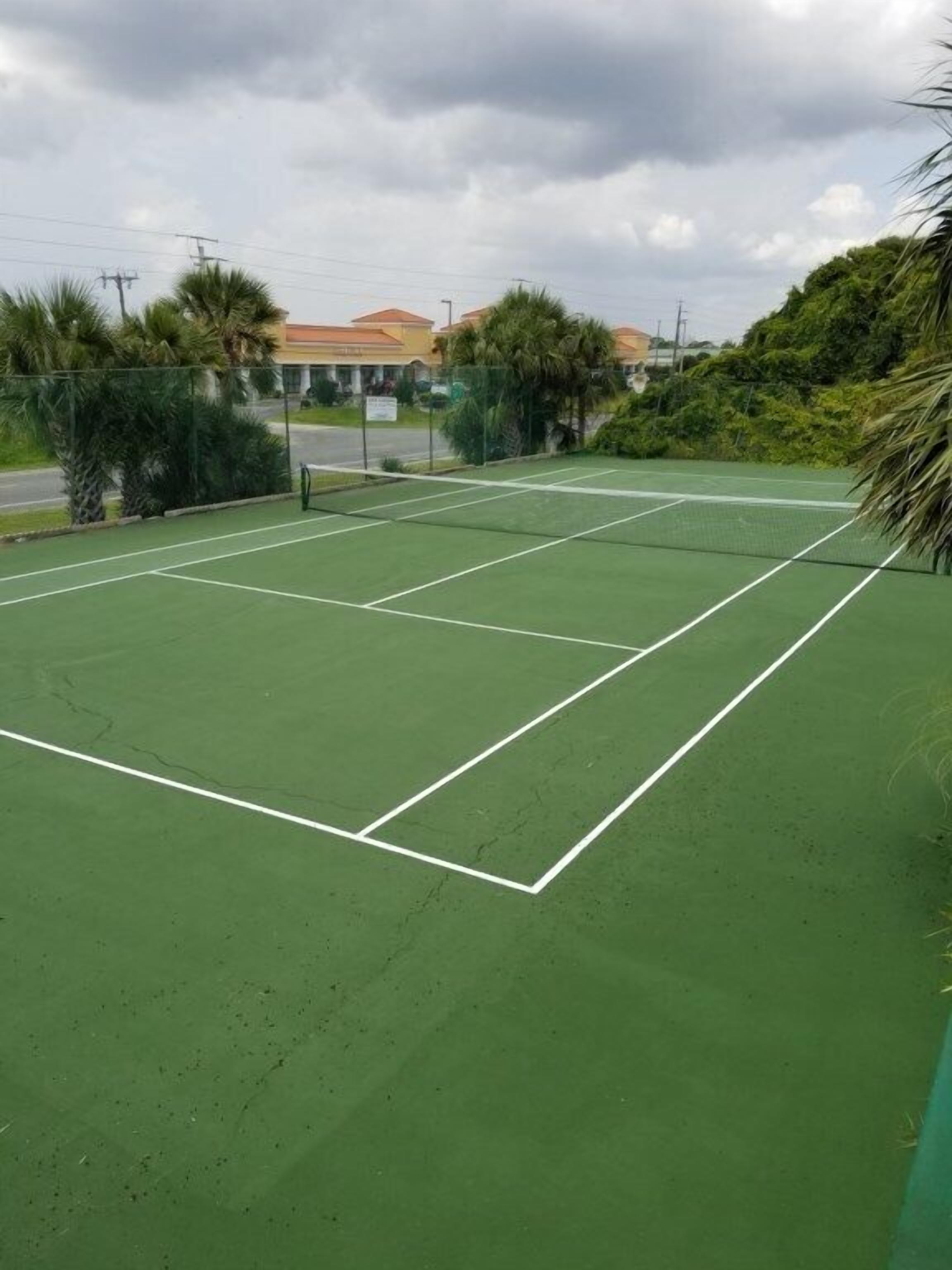 An outdoor tennis court with a green surface, surrounded by a fence and palm trees.