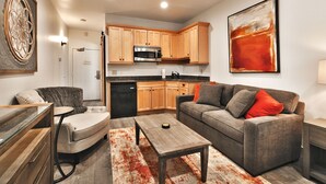 A modern living room with a grey sofa, a round coffee table, and a dining table with chairs. There is a kitchen area with wooden cabinets and a microwave. - Prospector Studio - Slps 4 (Park City)