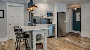 A modern kitchen with white cabinets, a stainless steel refrigerator, a microwave, and a sink. There are two bar stools and a hanging light fixture. - The Carlton* 3 min walk to Downtown Mt. Dora!!  (Mt Dora)
