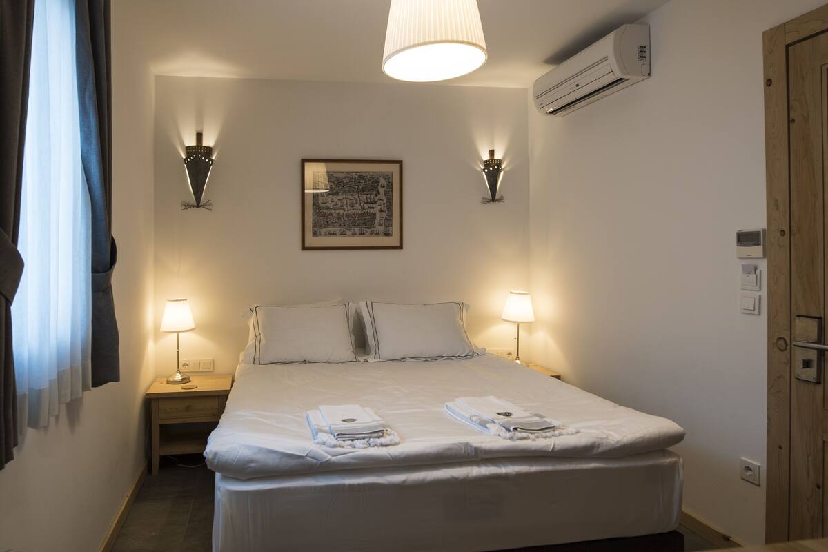 A neatly made bed with towels, two bedside lamps, a framed picture on the wall, and an air conditioning unit.