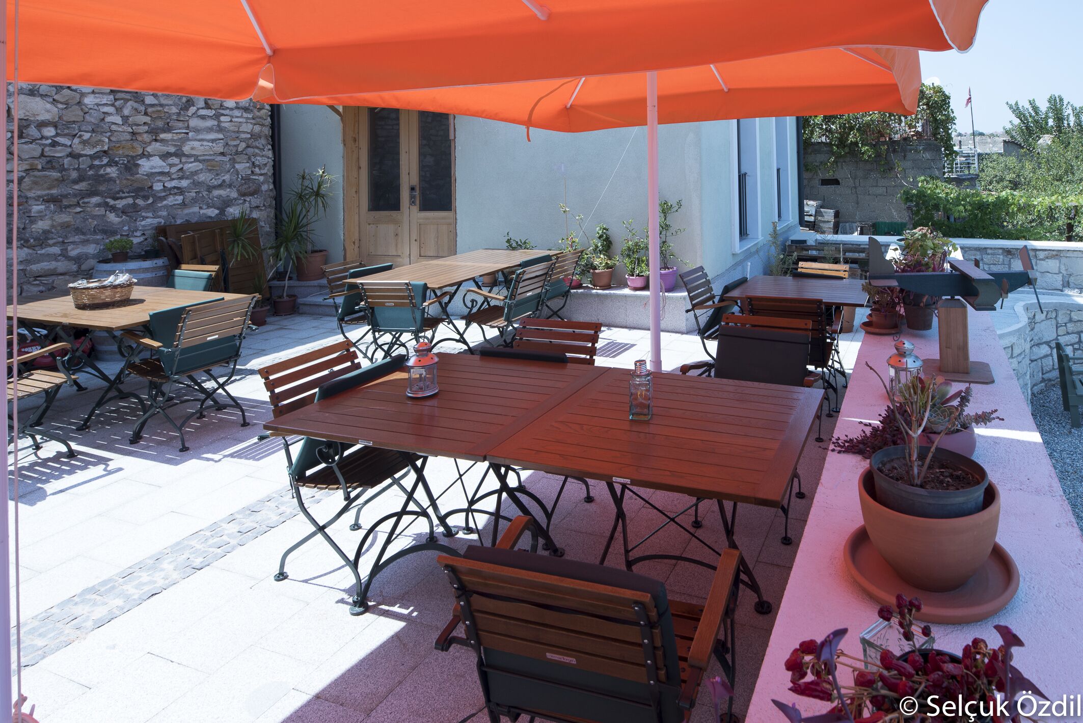 Un patio al aire libre con mesas y sillas de madera, plantas en macetas y un muro de piedra.