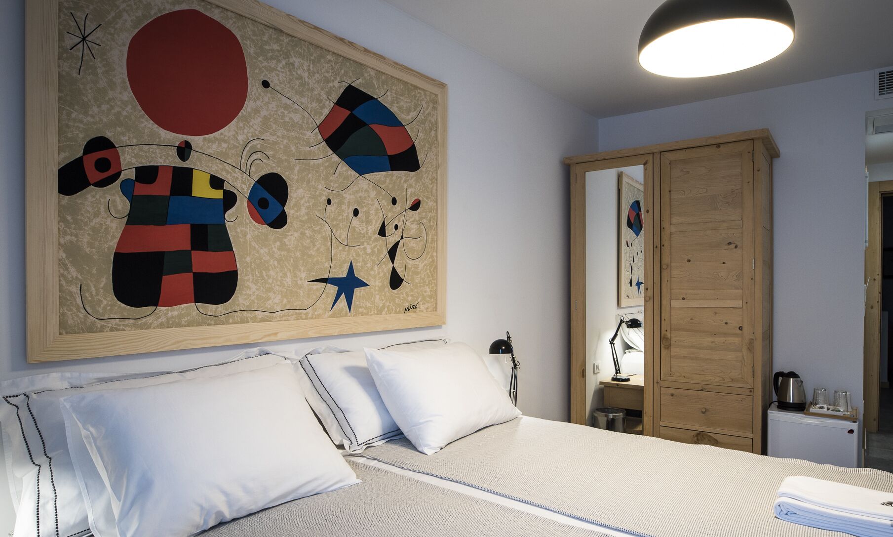 Un dormitorio con cama, almohadas blancas, un cuadro abstracto grande, un armario de madera y un escritorio con lámpara.