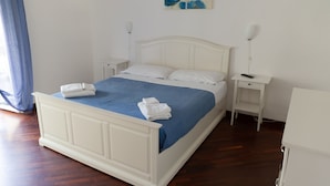 A neatly made bed with white linens and a blue bedspread, two bedside tables, a wall-mounted lamp, and a painting above the bed.