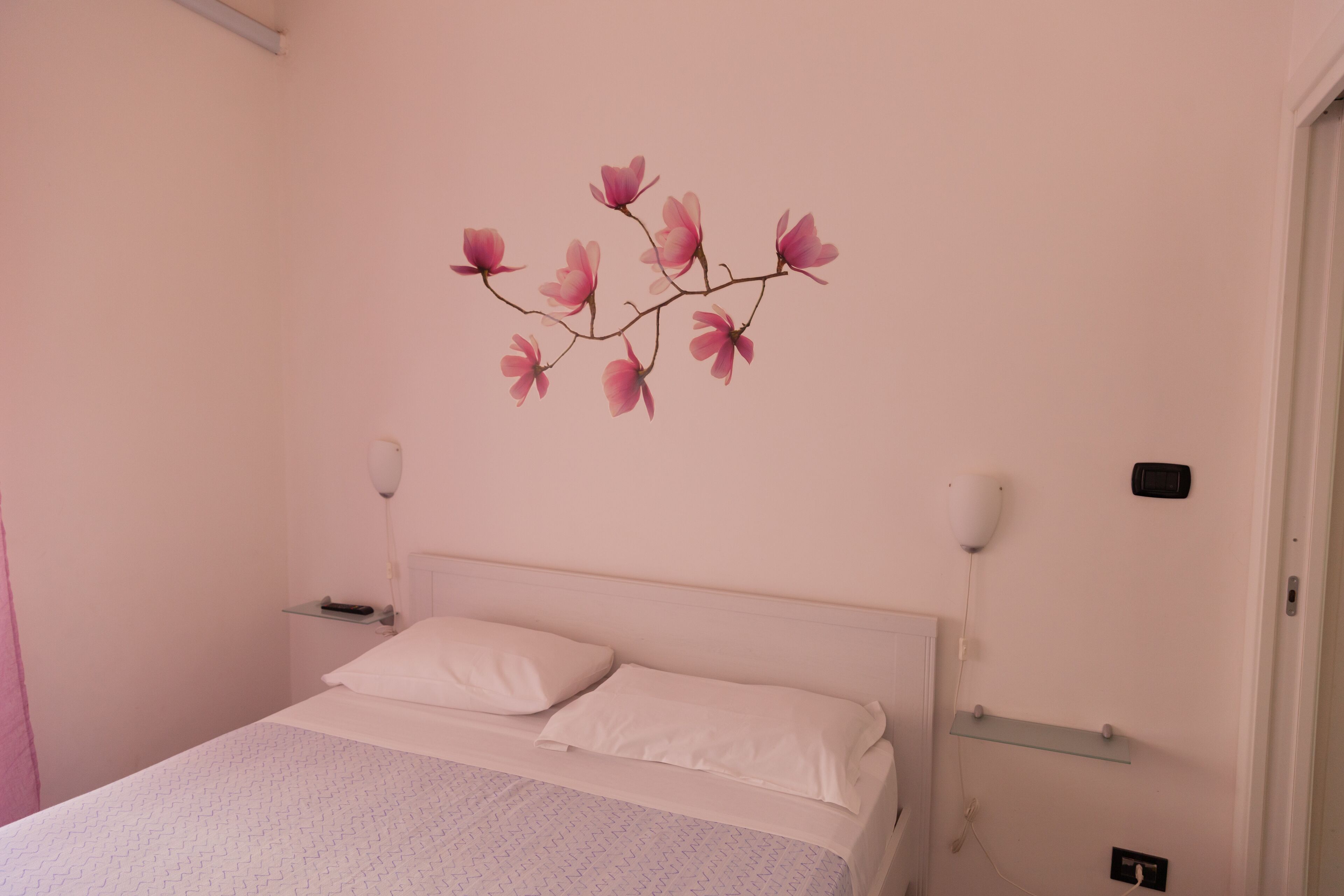 A bedroom with a bed, white bedding, a wall-mounted shelf, a bedside table, and a wall decoration of pink flowers.