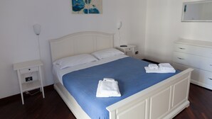 A neatly made bed with a blue bedspread and white pillows, a bedside table with a lamp, and a dresser with drawers.