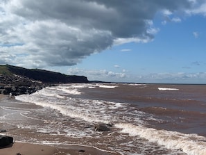 Une plage où les vagues se brisent sur le rivage, une falaise rocheuse et un ciel partiellement nuageux.