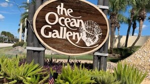 A wooden sign for 'The Ocean Gallery' with a seashell design, surrounded by plants and palm trees. - Ocean Gallery 26, Private Beach Access (St. Augustine)