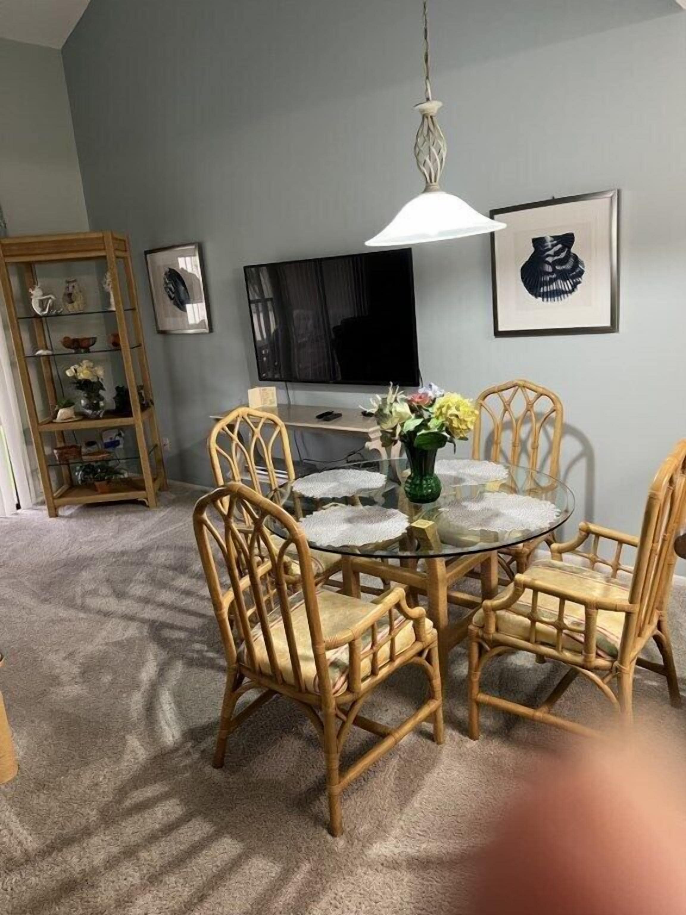 A dining area with a round glass table, four wicker chairs, a hanging light fixture, a television on the wall, and a bookshelf with decorative items.