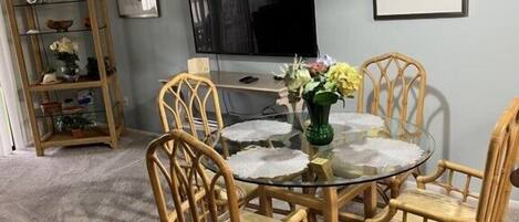 A dining area with a round glass table, four wicker chairs, a hanging light fixture, a television on the wall, and a bookshelf with decorative items.