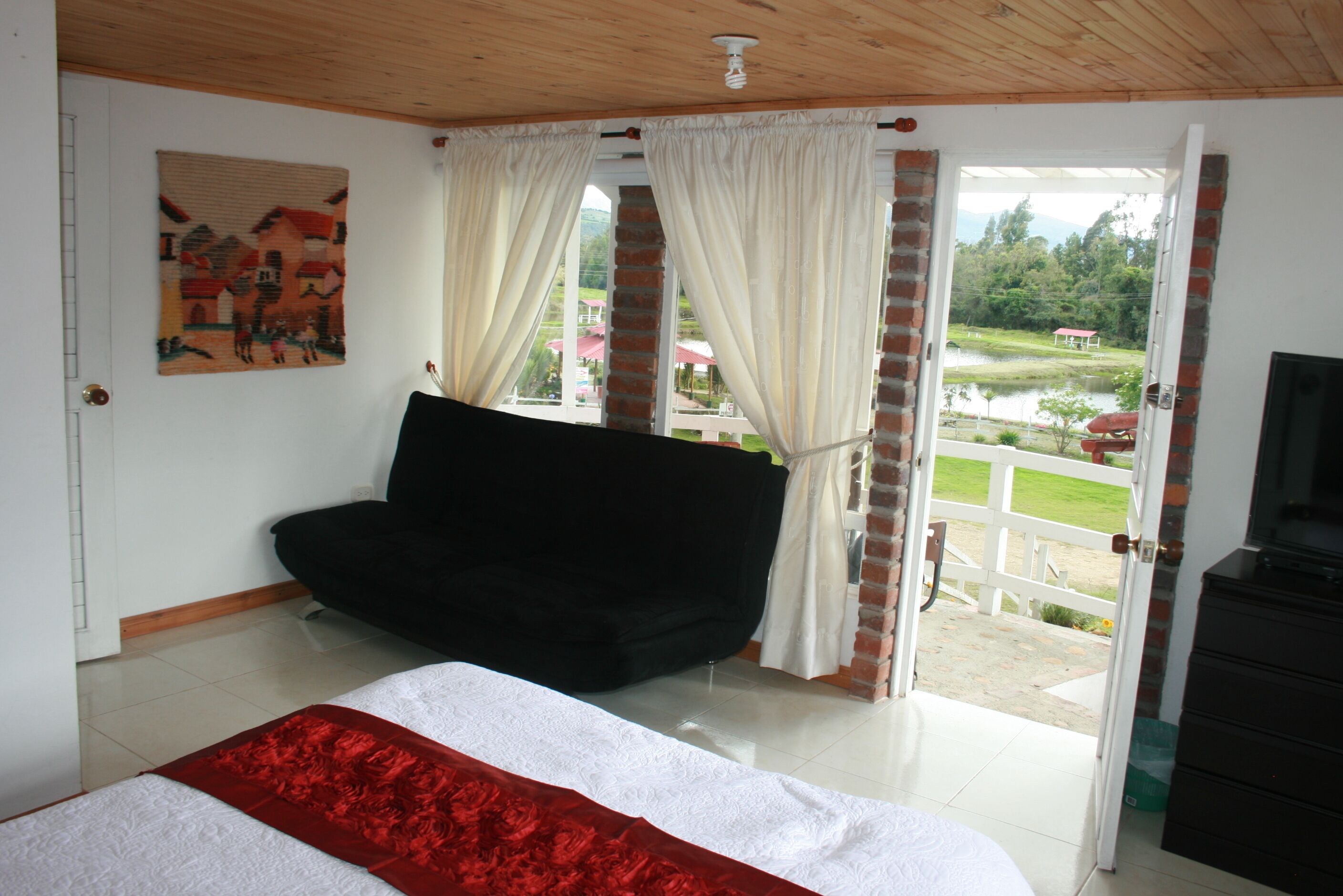 Un dormitorio con cama, sofá negro, un cuadro en la pared y vistas de un jardín y un edificio a través de una puerta.
