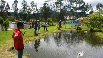 Un grupo de personas pescando en un estanque en un parque, con árboles y un parque infantil al fondo.