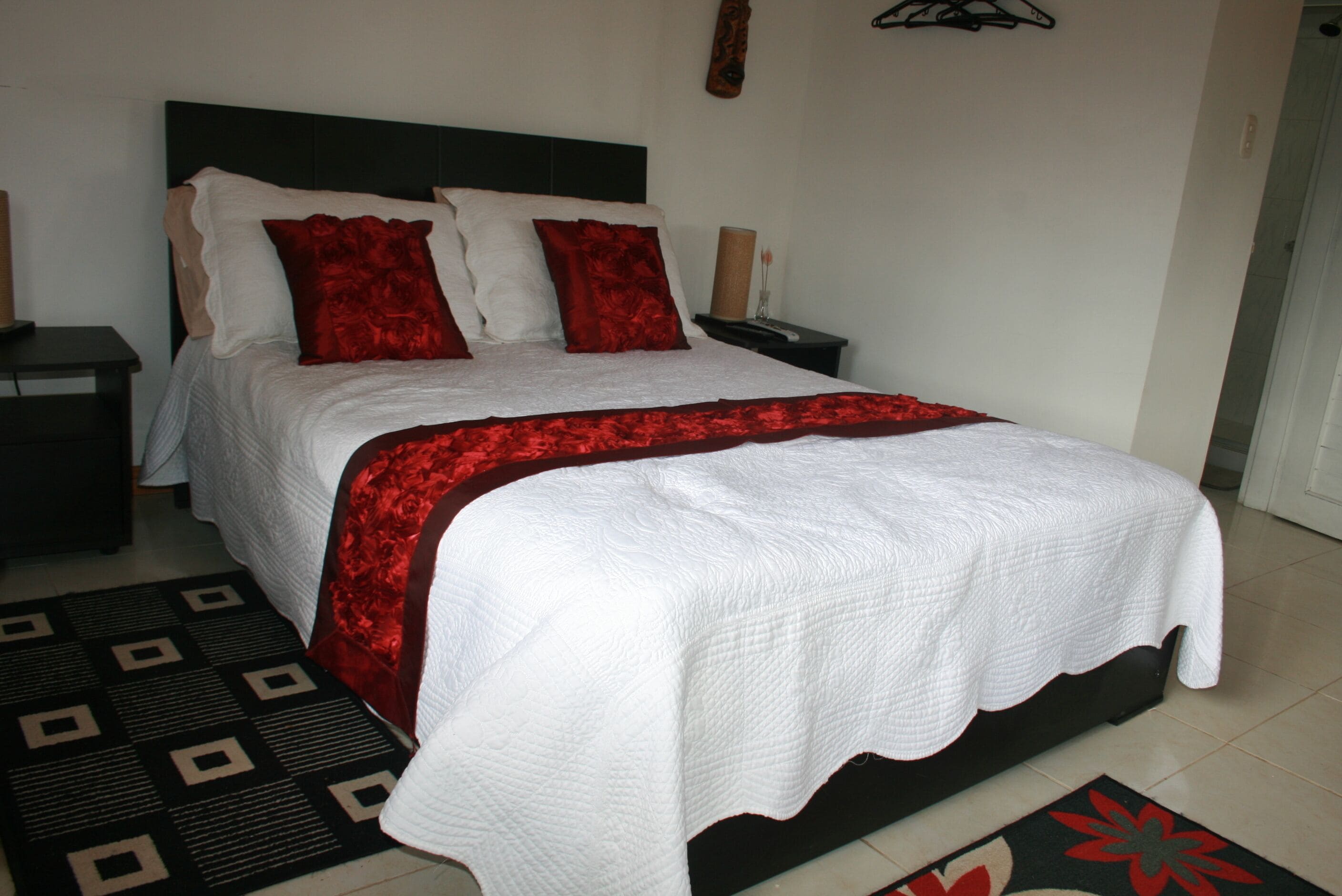 A neatly made bed with a white and red bedspread, a dark headboard, and a black and red patterned rug on the floor.