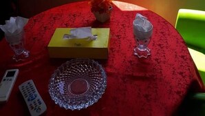 Uma mesa com toalha vermelha, um vaso com flores, uma caixa amarela, um controle remoto e um copo.